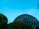 A geodesic dome structure is partially visible surrounded by trees under a clear blue sky. The geometric patterns on the dome are reflective, creating a mix of dark and light shades. There is a small bright object in the sky, possibly the moon.