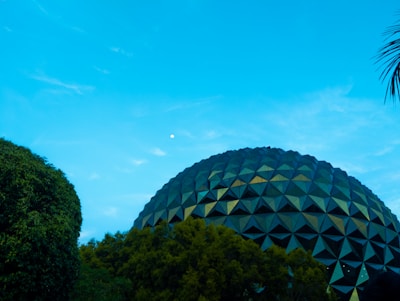 A geodesic dome structure is partially visible surrounded by trees under a clear blue sky. The geometric patterns on the dome are reflective, creating a mix of dark and light shades. There is a small bright object in the sky, possibly the moon.