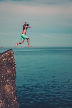 A person in turquoise swim shorts is captured mid-air after jumping off a rocky cliff into the calm sea below. The scene illustrates a sense of adventure and freedom. The clear blue water and sky provide a tranquil and serene backdrop.