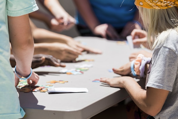 Several people are gathered around a table working together on a puzzle. Various hands are visible reaching for and placing pieces. The focus is on teamwork and collaboration.