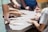 Several people are gathered around a table working together on a puzzle. Various hands are visible reaching for and placing pieces. The focus is on teamwork and collaboration.