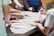 Several people are gathered around a table working together on a puzzle. Various hands are visible reaching for and placing pieces. The focus is on teamwork and collaboration.