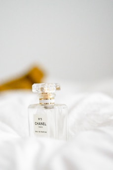 A clear bottle of Chanel No. 5 perfume rests on a soft white surface, with its elegant, minimalist design and gold accents capturing a sense of luxury. A blurred brown object in the background adds depth to the composition.