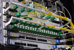 A close-up view of a network server rack filled with multiple fiber optic cables and connectors. The cables are predominantly yellow and green, some labeled with tags, and they are connected to various ports and modules. The setup appears highly organized and is likely part of a data center or telecommunications infrastructure.