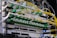 A close-up view of a network server rack filled with multiple fiber optic cables and connectors. The cables are predominantly yellow and green, some labeled with tags, and they are connected to various ports and modules. The setup appears highly organized and is likely part of a data center or telecommunications infrastructure.