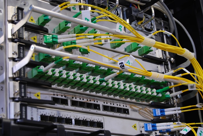 A close-up view of a network server rack filled with multiple fiber optic cables and connectors. The cables are predominantly yellow and green, some labeled with tags, and they are connected to various ports and modules. The setup appears highly organized and is likely part of a data center or telecommunications infrastructure.