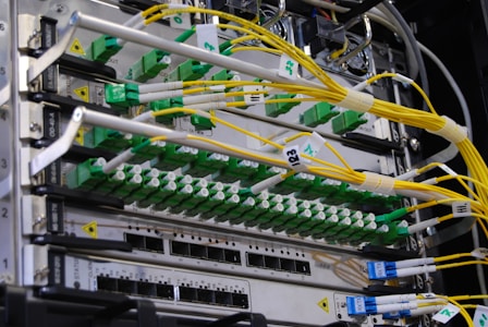 A close-up view of a network server rack filled with multiple fiber optic cables and connectors. The cables are predominantly yellow and green, some labeled with tags, and they are connected to various ports and modules. The setup appears highly organized and is likely part of a data center or telecommunications infrastructure.