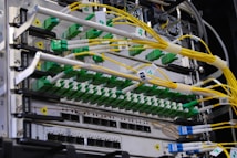 A close-up view of a network server rack filled with multiple fiber optic cables and connectors. The cables are predominantly yellow and green, some labeled with tags, and they are connected to various ports and modules. The setup appears highly organized and is likely part of a data center or telecommunications infrastructure.