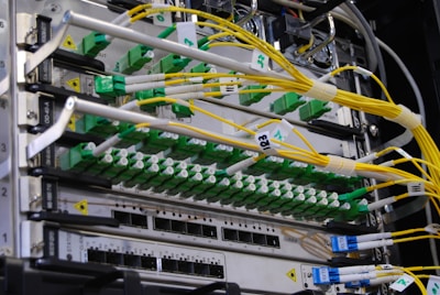 A close-up view of a network server rack filled with multiple fiber optic cables and connectors. The cables are predominantly yellow and green, some labeled with tags, and they are connected to various ports and modules. The setup appears highly organized and is likely part of a data center or telecommunications infrastructure.