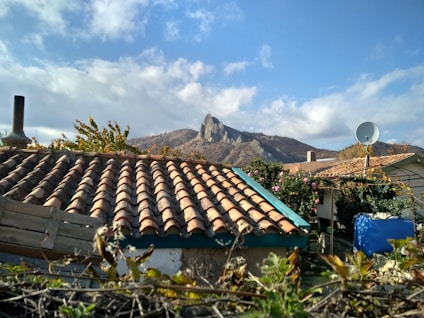 A rustic house with a tiled roof topped by a chimney and surrounded by foliage. In the background, a steep rocky mountain rises under a partly cloudy sky.