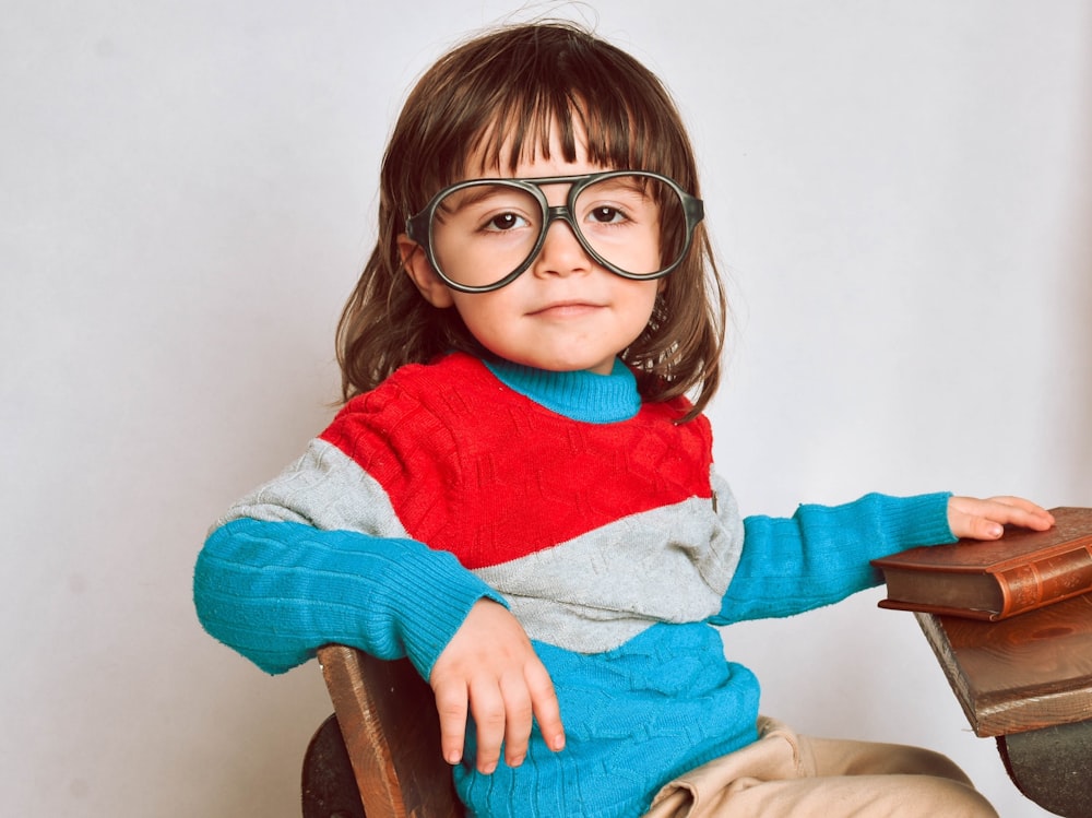 Mädchen mit rot gerahmter Brille und grauem Pullover sitzt auf braunem Holzstuhl