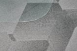 A close-up view of a textured, patterned carpet with varying shades of gray and a portion of a glass surface overlapping on the top left corner. The fibers of the carpet and the geometric design create a dynamic visual effect.