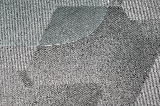 A close-up view of a textured, patterned carpet with varying shades of gray and a portion of a glass surface overlapping on the top left corner. The fibers of the carpet and the geometric design create a dynamic visual effect.