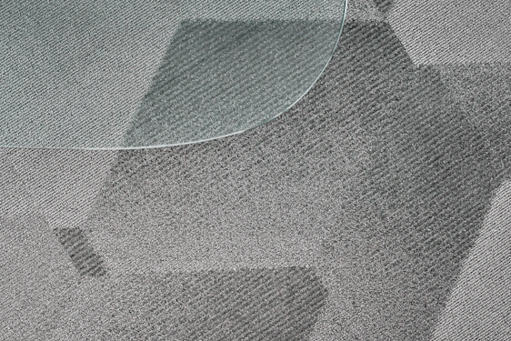 A close-up view of a textured, patterned carpet with varying shades of gray and a portion of a glass surface overlapping on the top left corner. The fibers of the carpet and the geometric design create a dynamic visual effect.