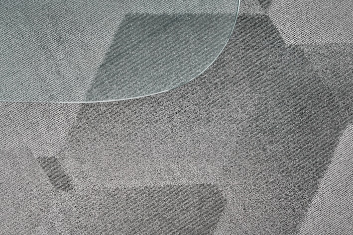 A close-up view of a textured, patterned carpet with varying shades of gray and a portion of a glass surface overlapping on the top left corner. The fibers of the carpet and the geometric design create a dynamic visual effect.