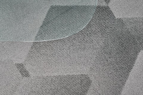 A close-up view of a textured, patterned carpet with varying shades of gray and a portion of a glass surface overlapping on the top left corner. The fibers of the carpet and the geometric design create a dynamic visual effect.