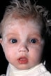 A close-up shot of a baby's face with white cream applied across the forehead, cheeks, and chin. The baby has wide blue eyes and a slightly open mouth, expressing a curious or surprised look. The hair is light, short, and somewhat spiky, with a dark background creating a strong contrast.