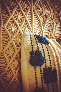A close-up view of a woven textile design and a cushion. The intricate pattern of the textile features a knotted and looped texture in a beige color. The cushion has a knitted cover with dark blue decorative patterns resembling flowers or dots, adding contrast to the beige background.
