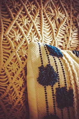 A close-up view of a woven textile design and a cushion. The intricate pattern of the textile features a knotted and looped texture in a beige color. The cushion has a knitted cover with dark blue decorative patterns resembling flowers or dots, adding contrast to the beige background.
