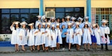 A group of graduates dressed in white gowns and caps are gathered outside a building with blue and yellow trim. They are standing in a joyful pose, some holding diplomas, and smiling.