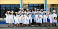 A group of graduates dressed in white gowns and caps are gathered outside a building with blue and yellow trim. They are standing in a joyful pose, some holding diplomas, and smiling.
