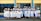A group of graduates dressed in white gowns and caps are gathered outside a building with blue and yellow trim. They are standing in a joyful pose, some holding diplomas, and smiling.