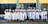A group of graduates dressed in white gowns and caps are gathered outside a building with blue and yellow trim. They are standing in a joyful pose, some holding diplomas, and smiling.