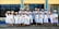 A group of graduates dressed in white gowns and caps are gathered outside a building with blue and yellow trim. They are standing in a joyful pose, some holding diplomas, and smiling.