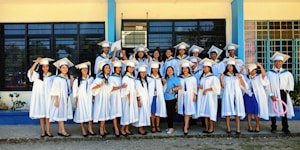 A group of graduates dressed in white gowns and caps are gathered outside a building with blue and yellow trim. They are standing in a joyful pose, some holding diplomas, and smiling.