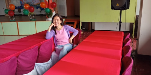 A woman is sitting comfortably on a row of red chairs with her elbow leaning on the back. She is smiling and wearing a purple jacket over a pink shirt with ripped jeans. Behind her, there are colorful balloons and a stage setup. A speaker is positioned to the right.