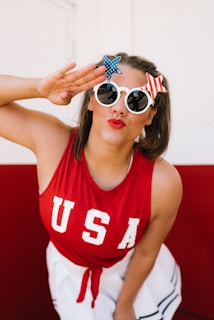 A person wearing a red tank top with 'USA' printed on it and white sunglasses adorned with star decorations. The scene is bright with a red and white background. The person has a playful expression with red lipstick and is posing with a salute gesture.