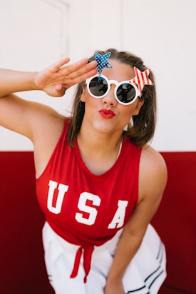 A person wearing a red tank top with 'USA' printed on it and white sunglasses adorned with star decorations. The scene is bright with a red and white background. The person has a playful expression with red lipstick and is posing with a salute gesture.