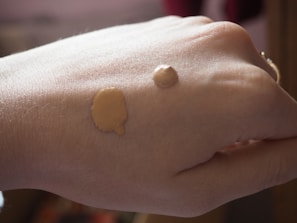 A close-up view of a hand with two different shades of liquid foundation applied on the back of it. The lighting is soft and natural, highlighting the texture of the skin and the makeup.