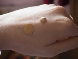 A close-up view of a hand with two different shades of liquid foundation applied on the back of it. The lighting is soft and natural, highlighting the texture of the skin and the makeup.