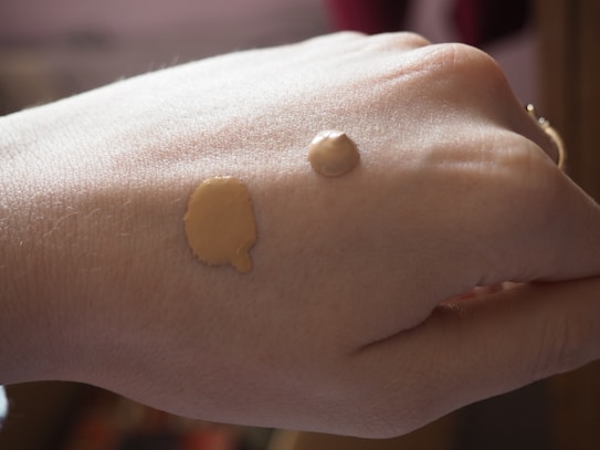 A close-up view of a hand with two different shades of liquid foundation applied on the back of it. The lighting is soft and natural, highlighting the texture of the skin and the makeup.