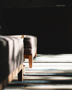 A close-up of wooden furniture legs cast in dramatic sunlight, creating distinct shadows on a striped, textured floor. The composition emphasizes light and shadow play with a focus on texture.