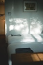Sunlight filters through a window, casting soft, dappled shadows on a neatly made bed. A neatly folded towel rests at the foot of the bed. A painting hangs on the light-colored wall, adding an artistic touch to the minimalistic decor. The scene conveys tranquility and simplicity.
