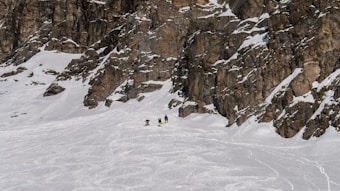 A snow-covered mountain landscape with rugged rocky cliffs. Three people are trekking through the snow towards the base of the cliffs, creating a sense of scale against the vastness of the environment. The snow is slightly disturbed by previous tracks, indicating a popular spot for outdoor activities.
