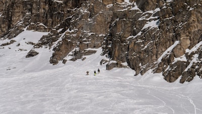 A snow-covered mountain landscape with rugged rocky cliffs. Three people are trekking through the snow towards the base of the cliffs, creating a sense of scale against the vastness of the environment. The snow is slightly disturbed by previous tracks, indicating a popular spot for outdoor activities.