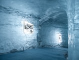A room carved out of ice with smooth, translucent walls, featuring illuminated frames displaying images. The floor is ice-covered, and the lighting gives a soft blue hue to the entire space. Electrical cords are visible running along the floor and walls, suggesting they power the illuminated displays.