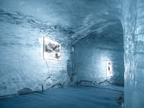 A room carved out of ice with smooth, translucent walls, featuring illuminated frames displaying images. The floor is ice-covered, and the lighting gives a soft blue hue to the entire space. Electrical cords are visible running along the floor and walls, suggesting they power the illuminated displays.