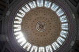 A large, ornate dome with intricate floral patterns and decorative designs. The dome features several glass panels allowing light to stream through, casting a bright glow. The structure appears to be part of an architectural building with elaborate detailing.