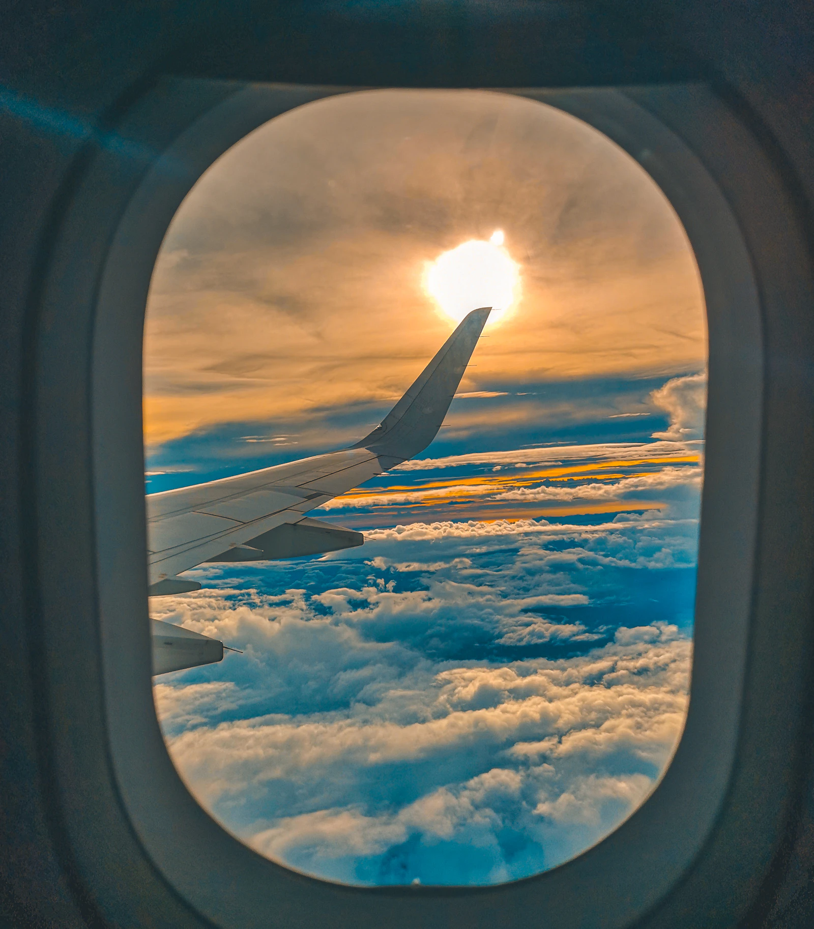 An airliner wing seen through a cabin window above a soft layer of clouds with the sun breaking through