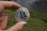A hand holds a silver coin featuring the Bitcoin logo against a blurred outdoor background with grass and a body of water.