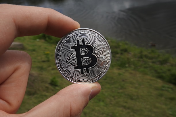 A hand holds a silver coin featuring the Bitcoin logo against a blurred outdoor background with grass and a body of water.