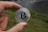 A hand holds a silver coin featuring the Bitcoin logo against a blurred outdoor background with grass and a body of water.