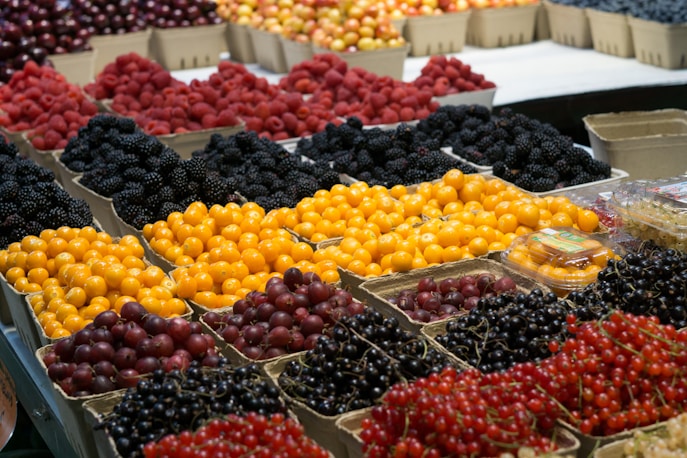 A variety of fresh fruits are displayed in cardboard boxes at a market or grocery store. Bright yellow gooseberries, dark purple grapes, red currants, blackberries, and raspberries are arranged in rows, creating a vibrant, colorful pattern.