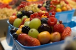 A variety of fresh fruits arranged in blue containers, including grapes, strawberries, raspberries, blackberries, and red currants. The fruits appear to be ripe and vibrant, with the background showing a blurred array of similar fruits.