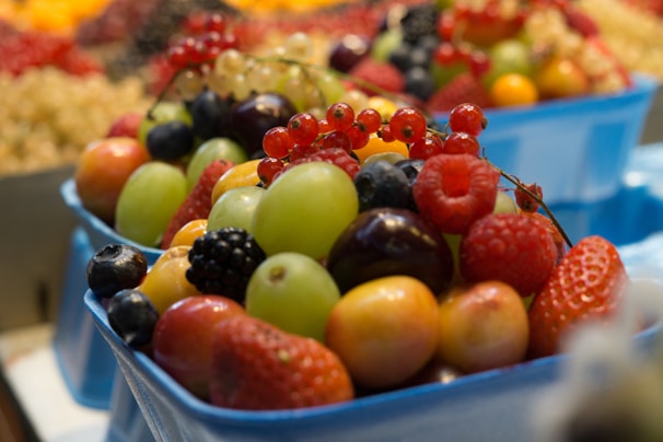 A variety of fresh fruits arranged in blue containers, including grapes, strawberries, raspberries, blackberries, and red currants. The fruits appear to be ripe and vibrant, with the background showing a blurred array of similar fruits.