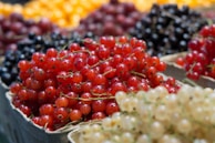 A close-up view of various colored berries, including red, black, and yellow, piled in small baskets. The foreground showcases vibrant red currants, while blurred background berries add depth to the image.